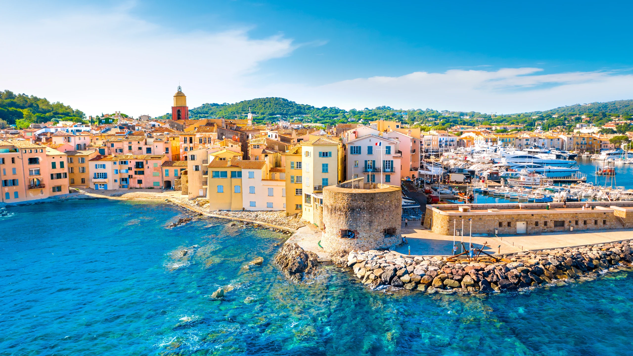 Luxury yacht anchored in the harbor of Saint-Tropez, France, with pastel-colored buildings, turquoise waters, and a lively waterfront in the background