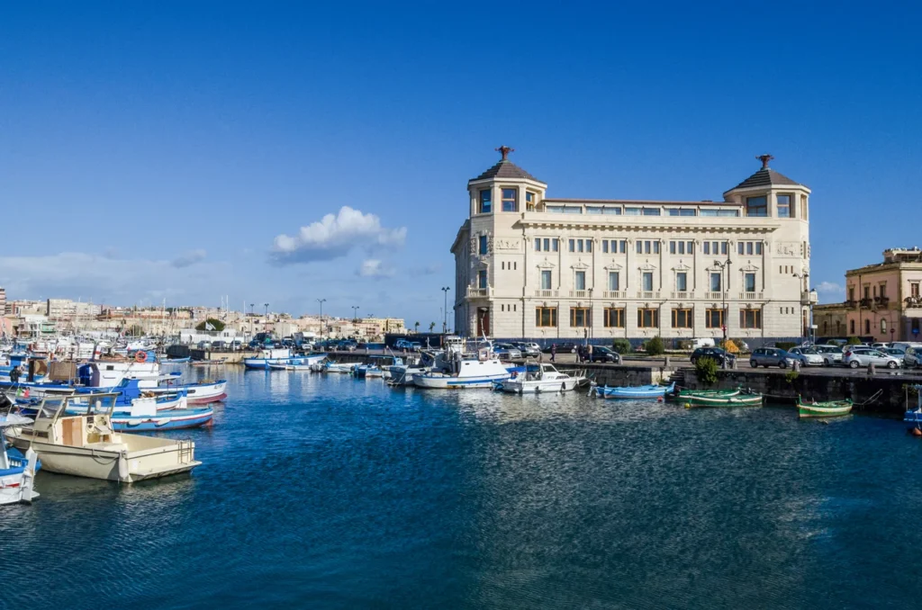 Luxury yacht anchored near the historic harbor of Syracuse, Sicily, Italy, with ancient architecture, turquoise waters, and a vibrant cityscape in the background.