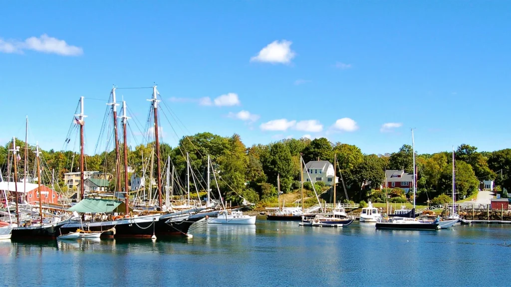 Luxury yacht anchored near a historic lighthouse in Maine, with snow-capped mountains, calm waters, and a rugged coastline in the background