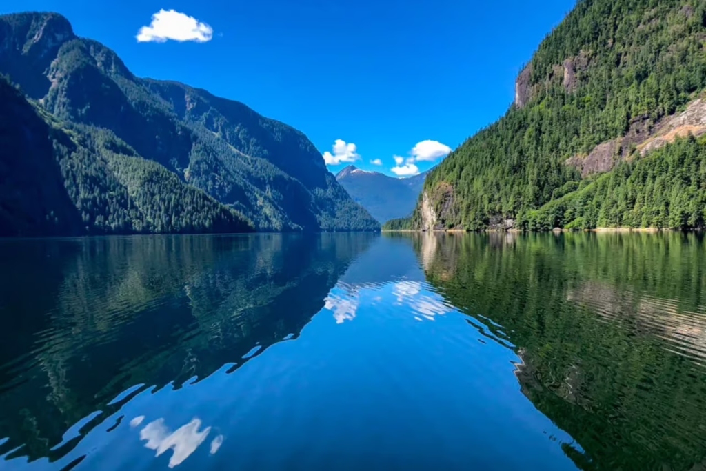 Luxury yacht cruising through a fjord-like waterway in Canada, surrounded by towering snowy mountain peaks and crystal-clear blue waters